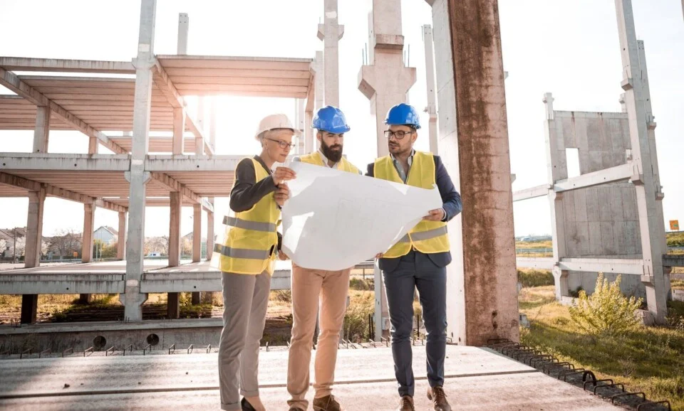 Photo d'un groupe composé d'une femme avec un casque blanc et des deux hommes avec un casque bleu tenant un plan devant un immeuble en construction.